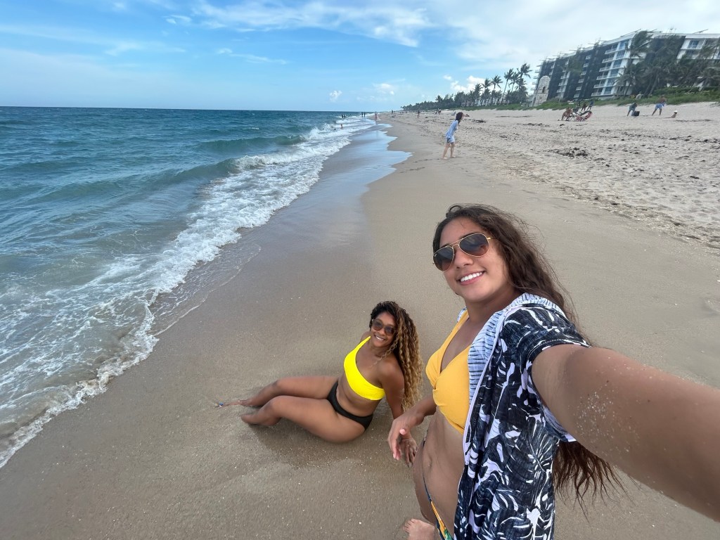 Two women in yellow swimwear enjoying a sunny day at the beach, one sitting on the sand and the other taking a selfie. Palm trees and a beachside building are visible in the background.