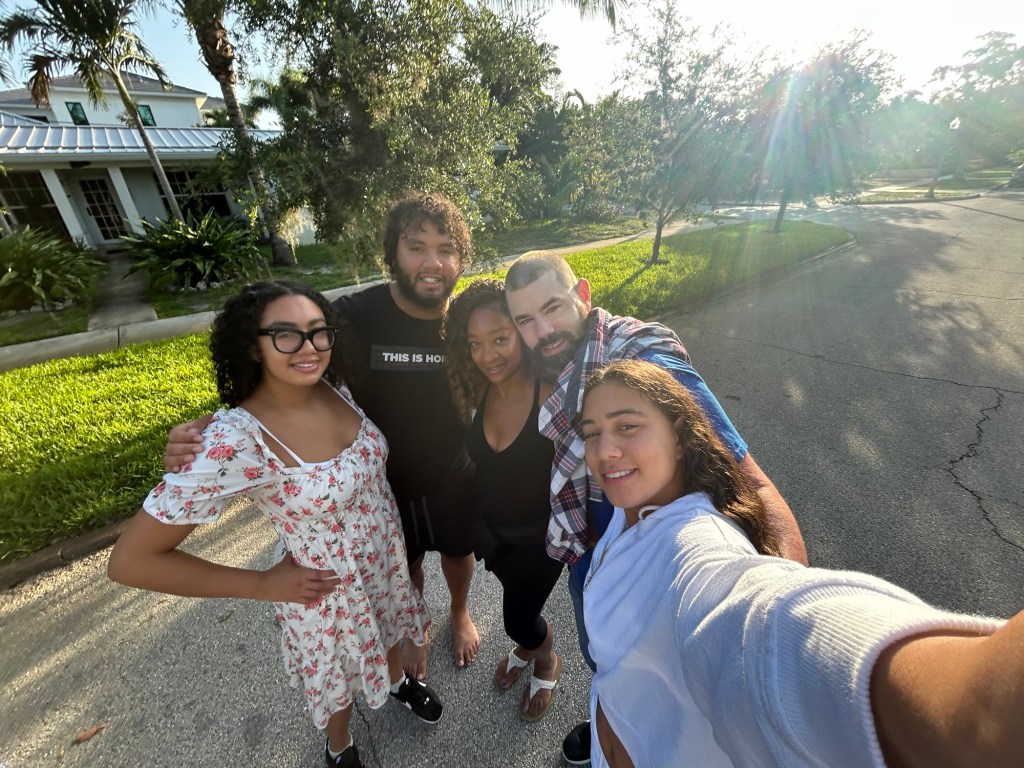 A group of five friends posing for a selfie outdoors, smiling and enjoying the sunlight in a green, suburban neighborhood.