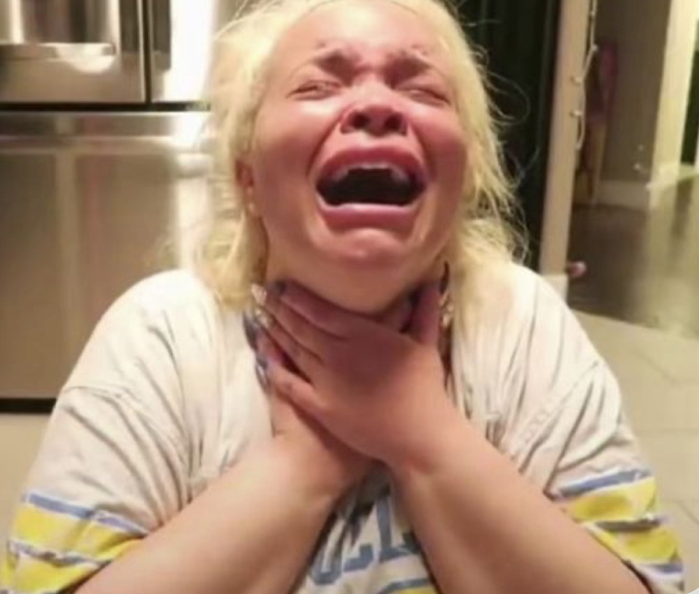 A person with blonde hair, wearing a oversized t-shirt, is sitting on the floor and crying while holding their throat in distress.